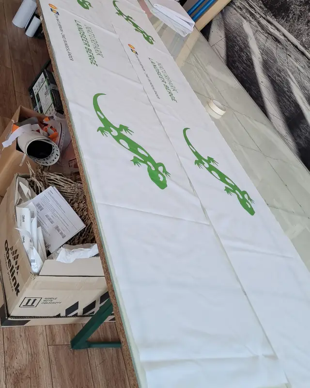 Bedruckte Stoffbanner mit grünem Eidechsen-Logo auf dem langen Werktisch
